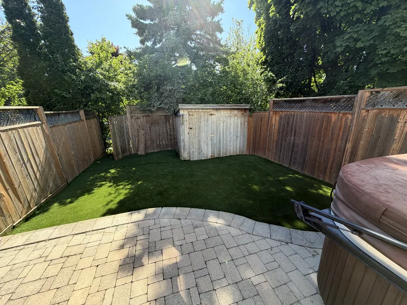 The same yard finished, artificial turf laid to the curve of the paver patio inside the same fence line.