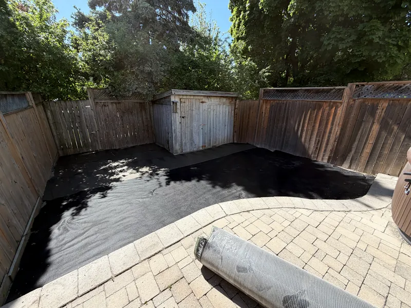 The same yard before: stripped to bare underlay with a roll of artificial turf waiting on the patio.